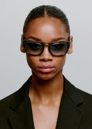Person wearing dark sunglasses and a black outfit against a neutral background