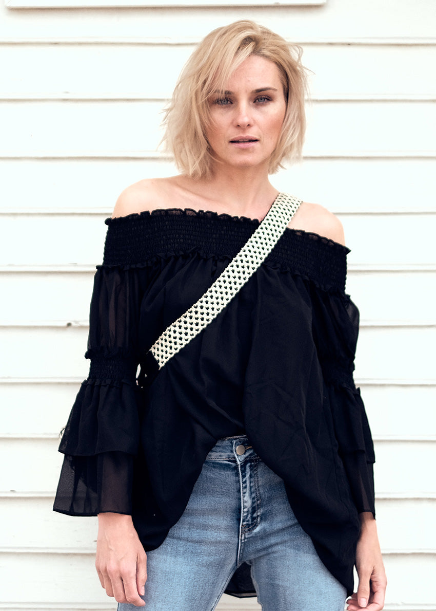 A woman wearing a black off-the-shoulder top with bracelet sleeves and tiered ruffle cuffs, paired with blue jeans and accessorized with a black purse with a checkered pattern.