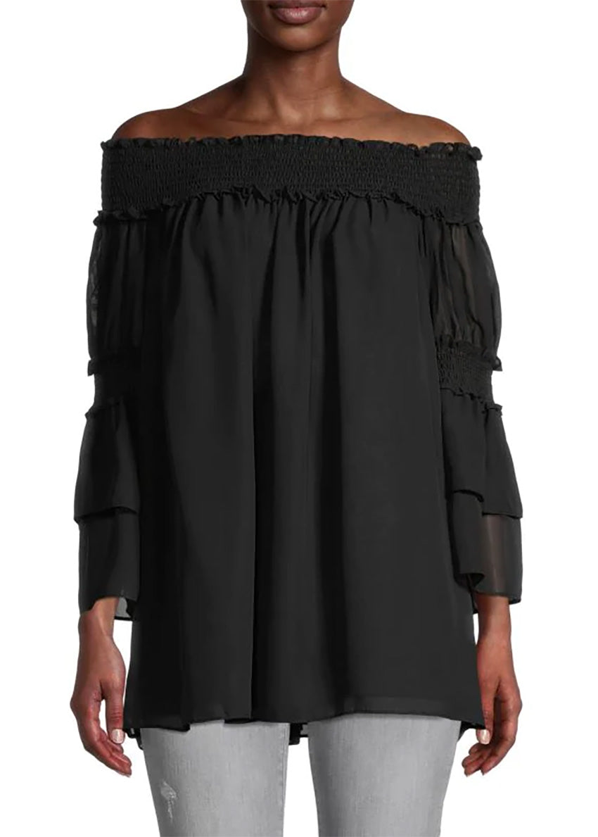 A woman wearing a black off-the-shoulder top with bracelet sleeves and tiered ruffle cuffs, paired with grey jeans