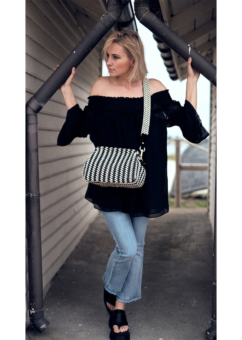 A woman wearing a black off-the-shoulder top with bracelet sleeves and tiered ruffle cuffs, paired with blue jeans and accessorized with a black purse with a checkered pattern.