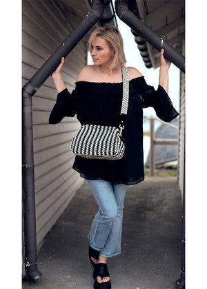 A woman wearing a black off-the-shoulder top with bracelet sleeves and tiered ruffle cuffs, paired with blue jeans and accessorized with a black purse with a checkered pattern.