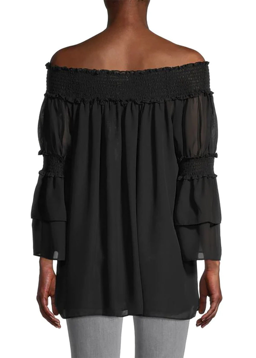 A woman wearing a black off-the-shoulder top with bracelet sleeves and tiered ruffle cuffs, paired with grey jeans 