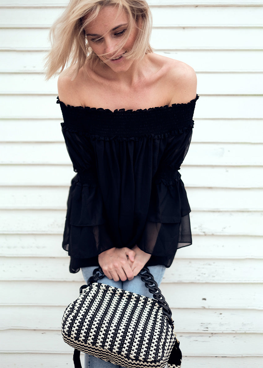 A woman wearing a black off-the-shoulder top with bracelet sleeves and tiered ruffle cuffs, paired with blue jeans and accessorized with a black purse with a checkered pattern.