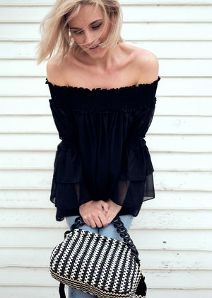 A woman wearing a black off-the-shoulder top with bracelet sleeves and tiered ruffle cuffs, paired with blue jeans and accessorized with a black purse with a checkered pattern.