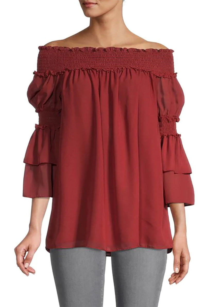 A woman wearing a red off-the-shoulder top with bracelet sleeves and tiered ruffle cuffs, paired with grey jeans