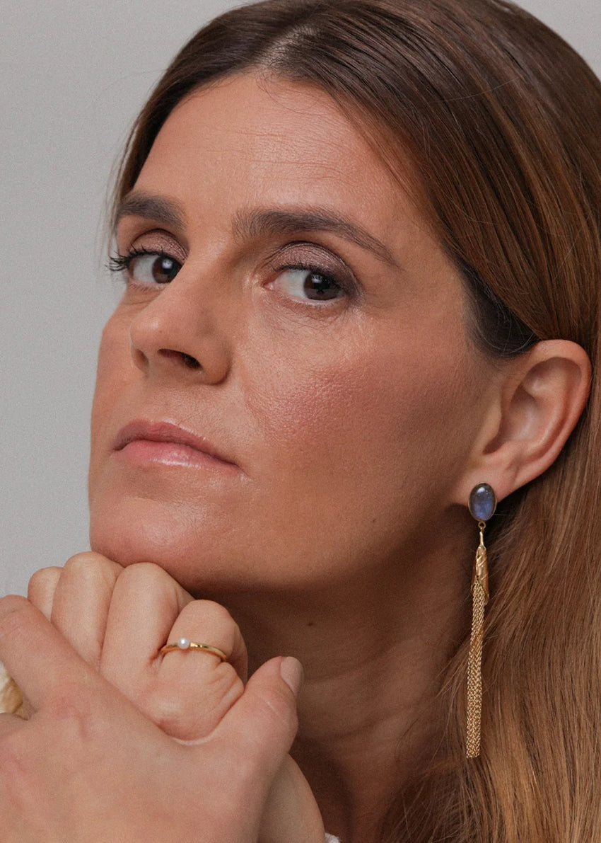 Woman with long brown hair wearing a gold earring and ring, against a neutral background