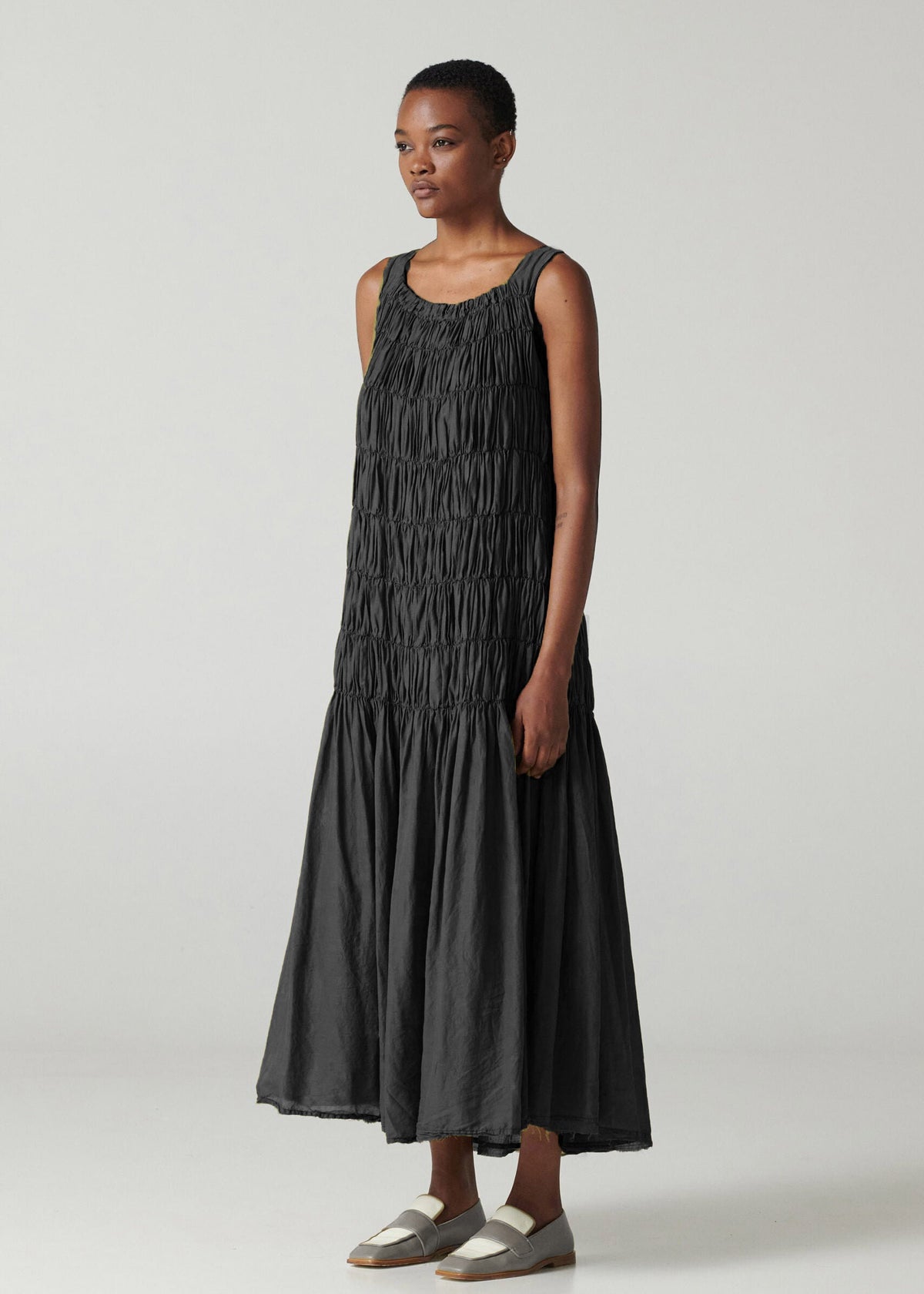 A woman standing wearing a long sleeveless black dress with square neck and wide straps, made of airy cotton voile fabric, and a shirred front.