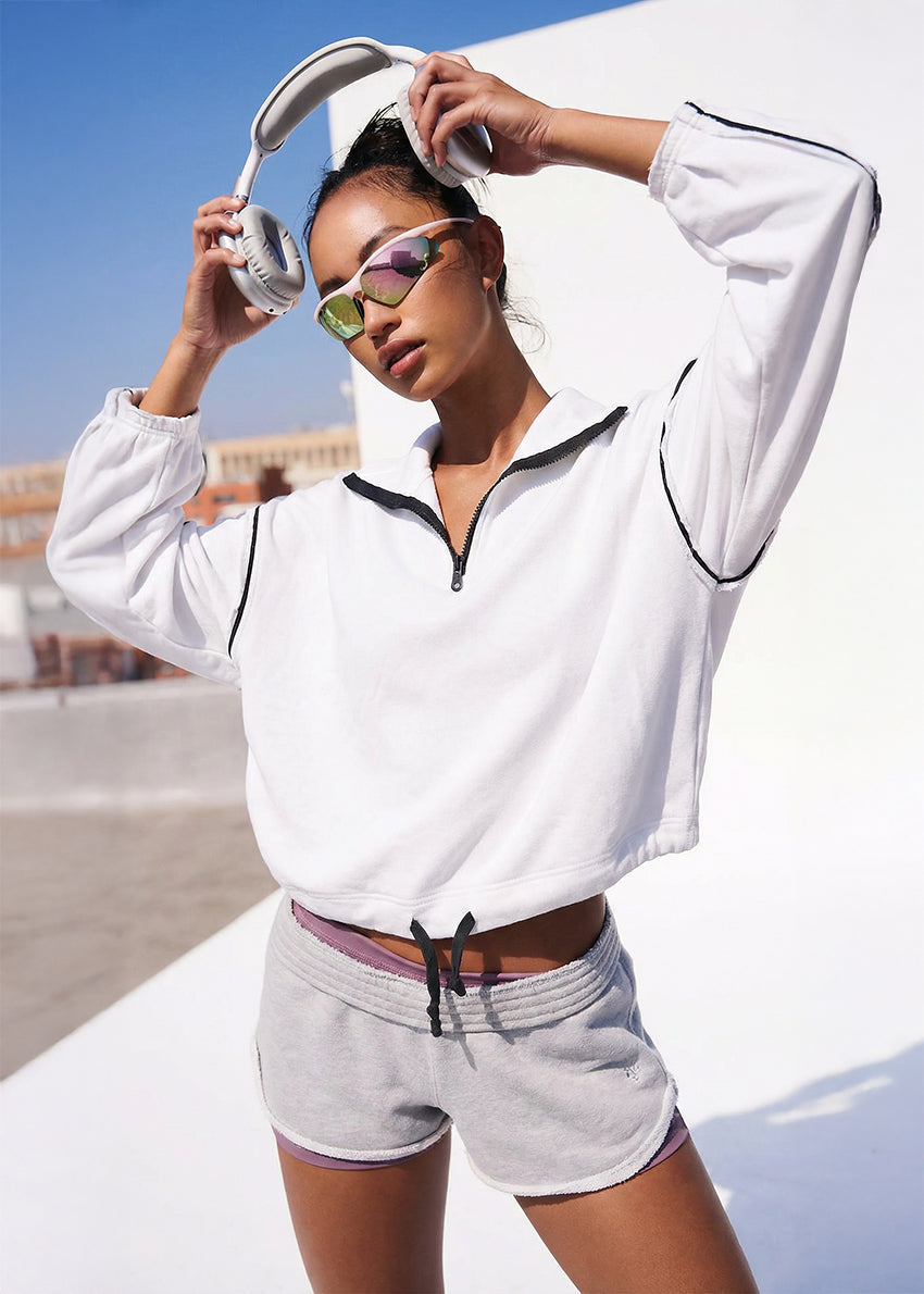 Woman wearing a white sweatshirt and gray shorts with sunglasses and headphones, standing on a white surface.