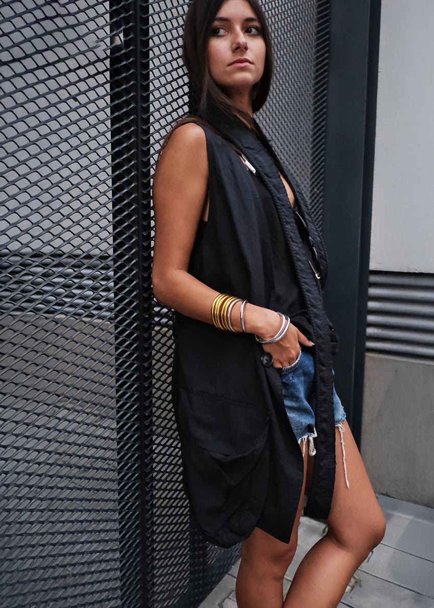 Woman wearing a black sleeveless vest and denim shorts leaning against a textured wall.
