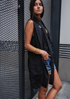 Woman wearing a black sleeveless vest and denim shorts leaning against a textured wall.