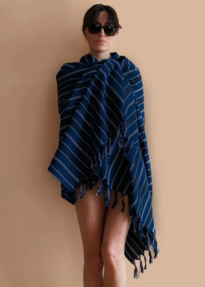 A person standing, wearing a blue striped towel with fringes, sunglasses, and showcasing a smile.