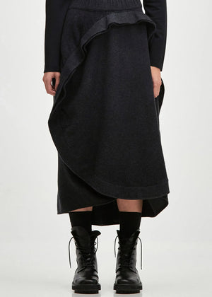 A person standing wearing an asymmetrical skirt with a ruffled design, in a dark color, with long sleeves and black boots.