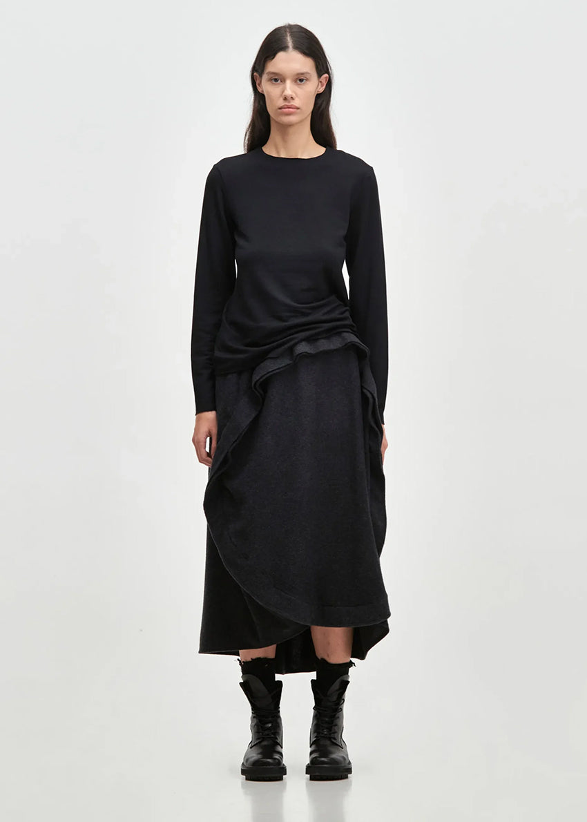 A person standing wearing an asymmetrical skirt with a ruffled design, in a dark color, with long sleeves and black boots.