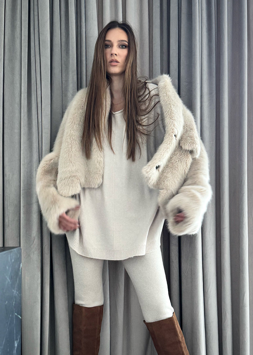 A woman wearing a plush faux fur coat with an open collar and front pockets, paired with white pants and brown boots.