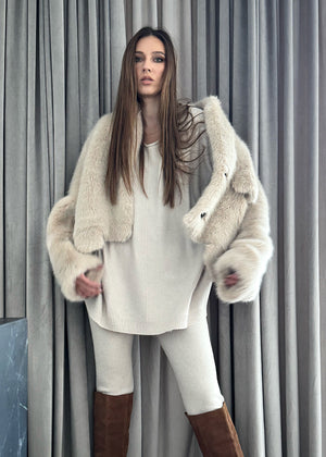 A woman wearing a plush faux fur coat with an open collar and front pockets, paired with white pants and brown boots.
