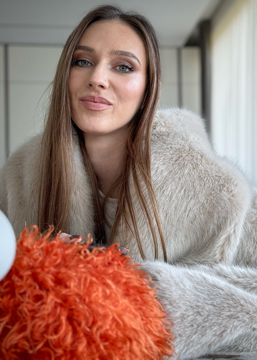 A woman sitting wearing a plush faux fur coat with an open collar and front pockets, paired with white pants and brown boots.