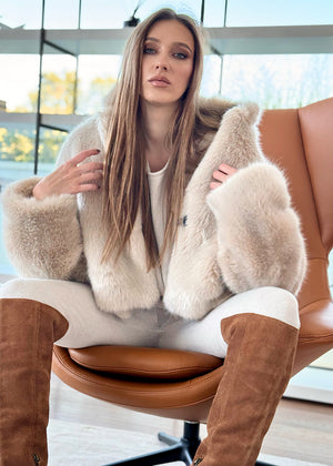 A woman sitting wearing a plush faux fur coat with an open collar and front pockets, paired with white pants and brown boots.