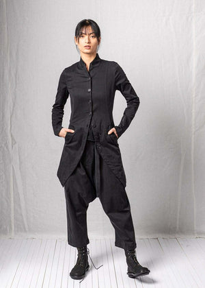 Person wearing a black outfit with a long jacket and high-waisted pants against a plain background