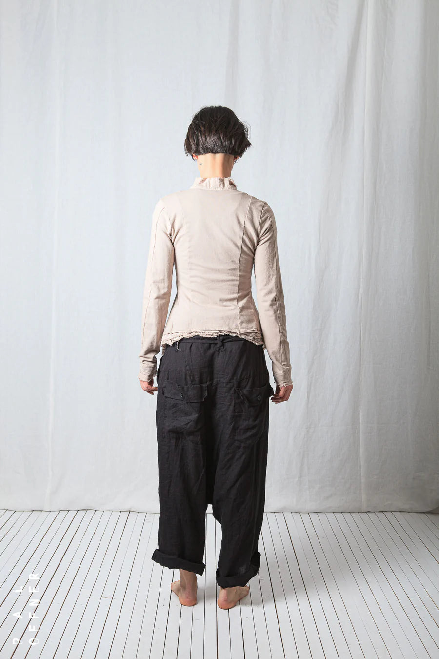 Person wearing a beige jacket and black pants standing against a white curtain.