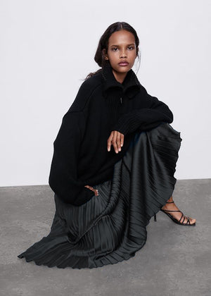 A person wearing a black pleated skirt and a black jacket, posing with one hand on their knee.