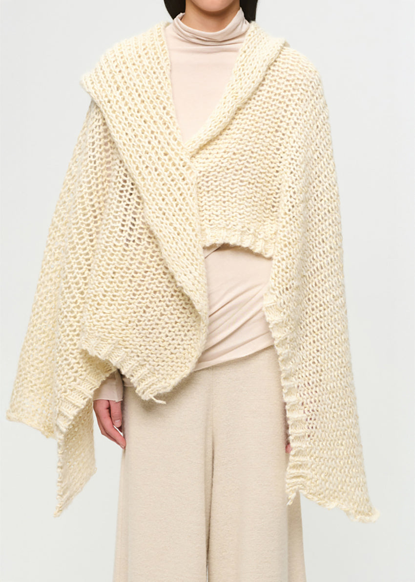 Person wearing a cream knitted shawl over a light outfit on a plain background