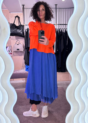 Woman taking a mirror selfie wearing an orange top and blue dress in a clothing store.