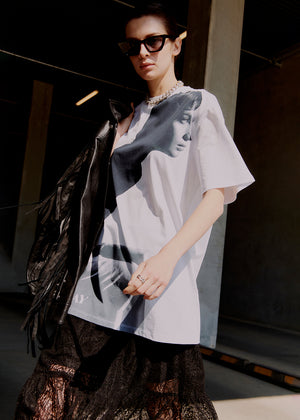 Woman wearing a white oversized shirt with a graphic design, standing in a dark indoor setting.