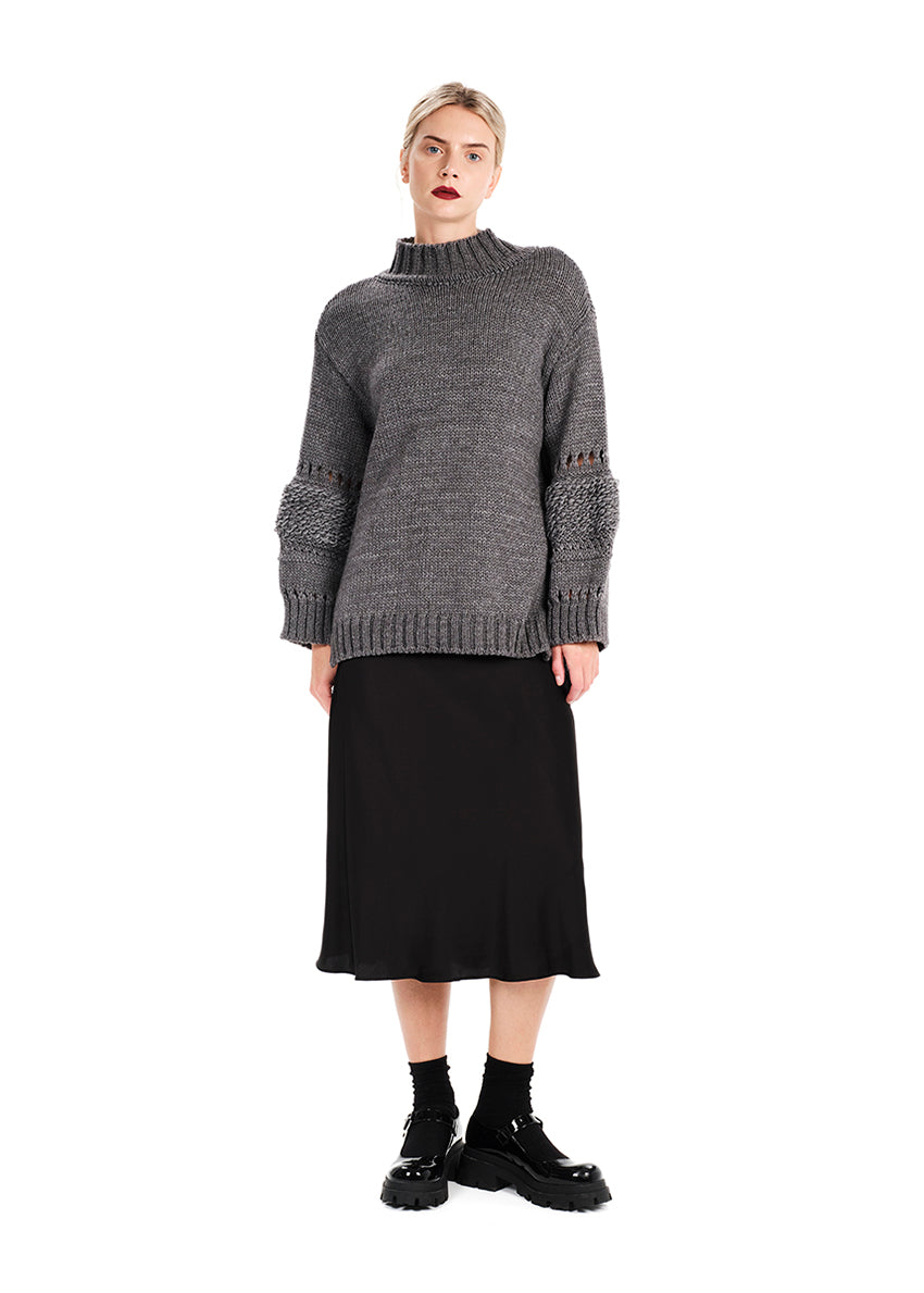 A model wearing a long-sleeved, knit sweater in a loose fit with bell sleeves and a ribbed hem, paired with a black skirt and black boots.