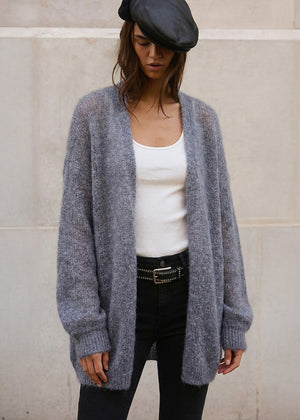 Person wearing a gray cardigan over a white top and black pants against a light-colored wall.