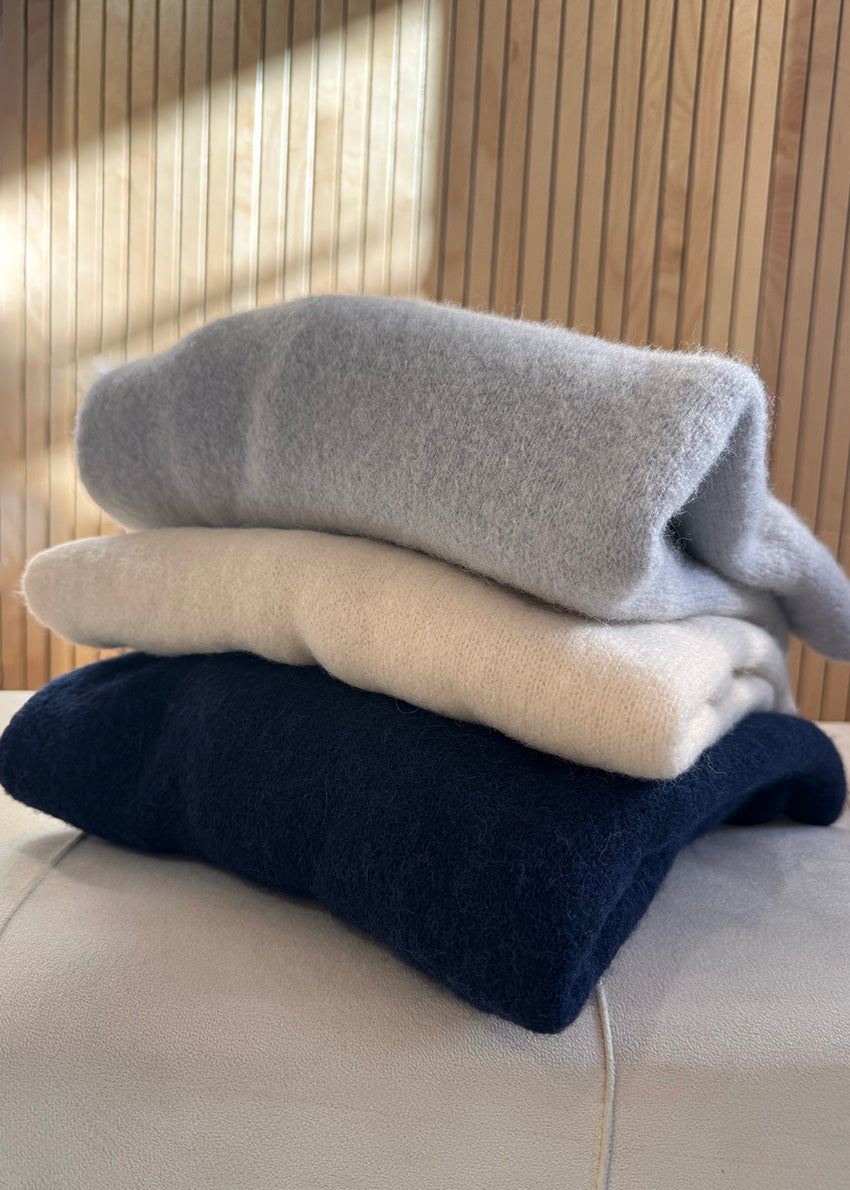 Stack of three folded sweaters in gray, white, and navy on a light surface with a wooden background.