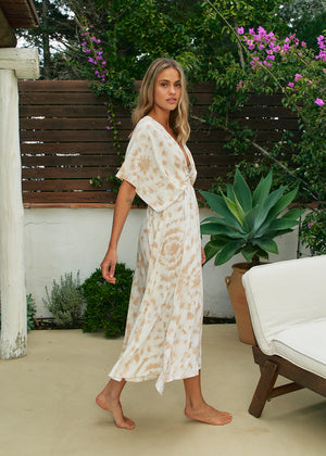 A woman wearing a bohemian style dress with a deep V-neckline and open batwing sleeves, featuring a beige and brown floral pattern. The dress is flowing and has a wrap front with an elasticated back waist.