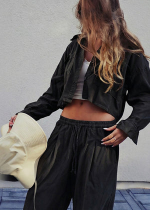 Person wearing a black jacket and pants holding a beige hat against a light gray wall.