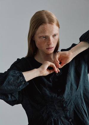 A person with red hair is seen from the back, adjusting the collar of a long-sleeved, black, loose-fit shirt.