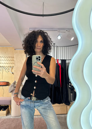 Person taking a mirror selfie wearing a black sleeveless vest and light blue jeans in a store setting.