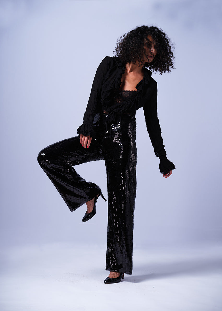 A person wearing black wide-leg trousers embellished with sequins, paired with a black top and black shoes. The trousers have a high waist and are lined with viscose and spandex/elastane for stretch.