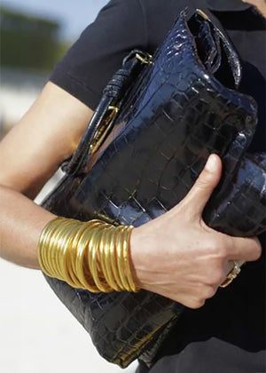 A person wearing a black leather bag and a 22Kt gold leaf bracelet.