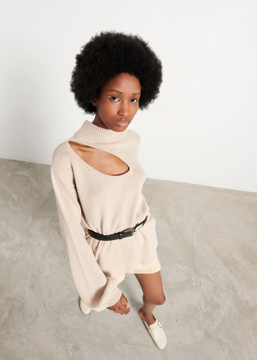 A woman wearing a beige, medium-length sweater with wide slits on the clavicle and shoulder sides, paired with a black belt.