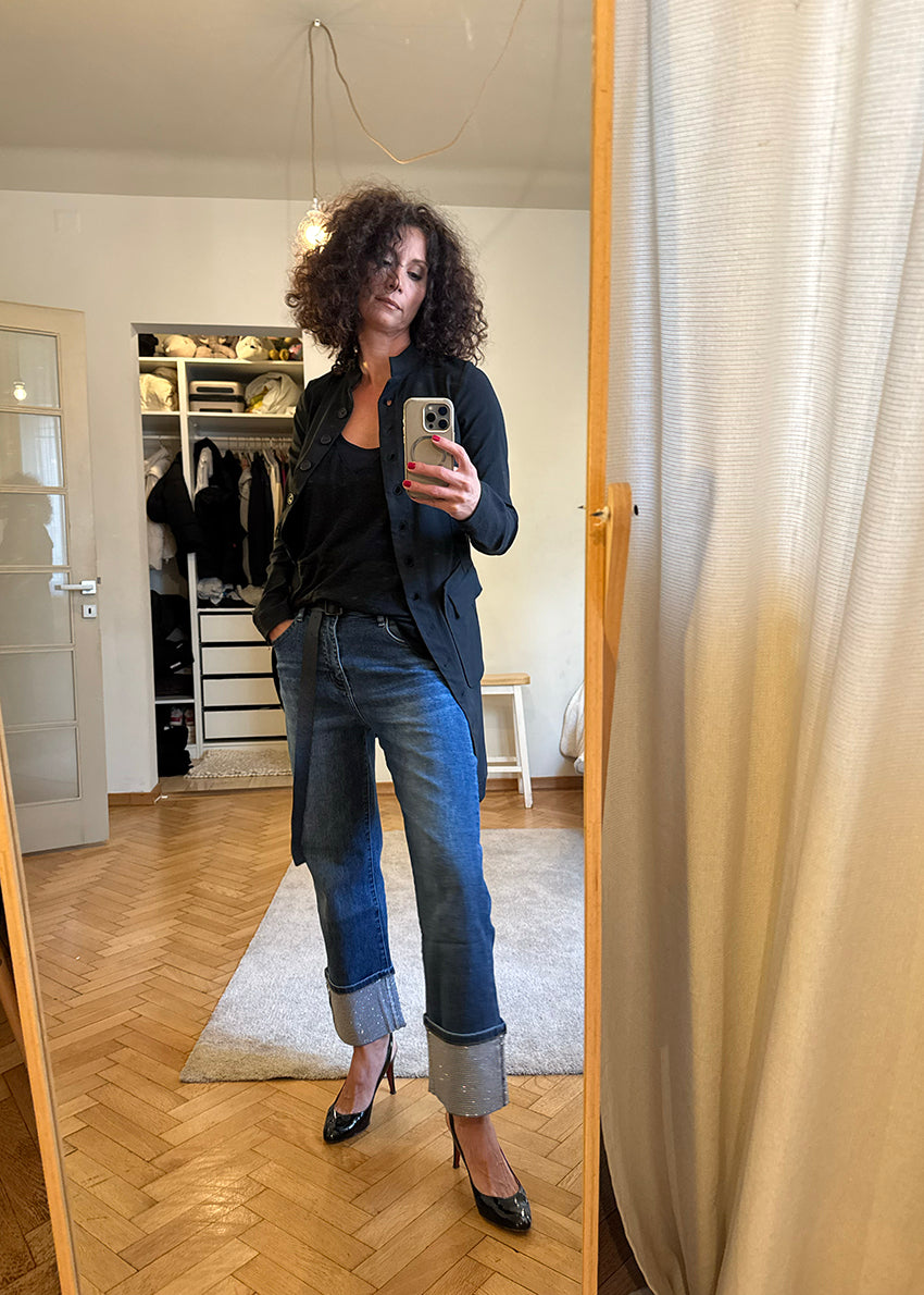 Woman taking a mirror selfie wearing a black jacket, black top, blue jeans, and black heels in a room with wooden floor and white walls.