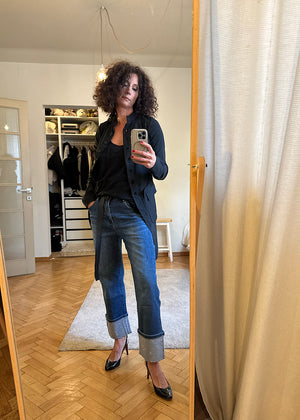 Woman taking a mirror selfie wearing a black jacket, black top, blue jeans, and black heels in a room with wooden floor and white walls.