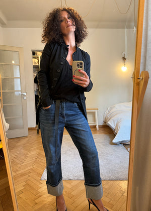 Woman taking a mirror selfie wearing a black jacket and blue jeans in a bedroom.