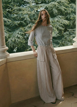 Woman in a light gray outfit standing on a balcony with greenery in the background