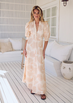A woman wearing a beige and white shell print maxi dress with 3/4 sleeves and a V-neck, standing on a deck.