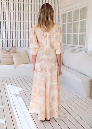 A woman wearing a beige and white shell print maxi dress with 3/4 sleeves and a V-neck, standing on a deck.
