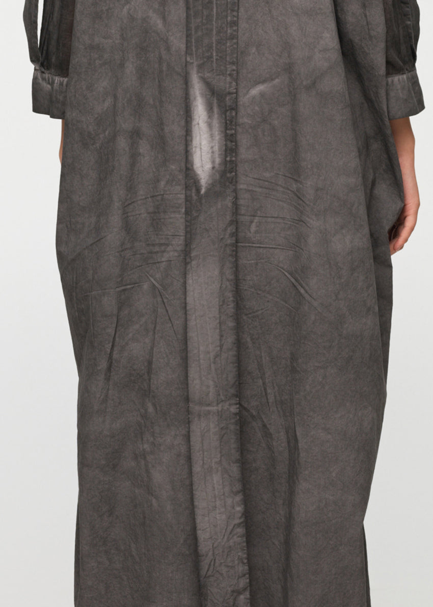 A person standing wearing a long, loose-fitting, grey shirt with 3/4-length sleeves, a collar, and buttons down the front. The shirt features a decorative vertical belt at the back and two side pockets.