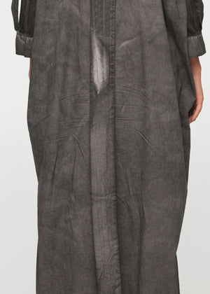 A person standing wearing a long, loose-fitting, grey shirt with 3/4-length sleeves, a collar, and buttons down the front. The shirt features a decorative vertical belt at the back and two side pockets.