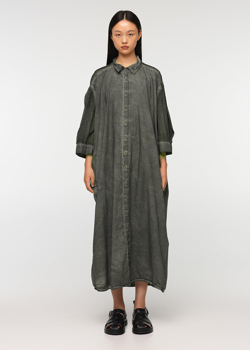 A person standing wearing a long, loose-fitting, grey shirt with 3/4-length sleeves, a collar, and buttons down the front. The shirt features a decorative vertical belt at the back and two side pockets.