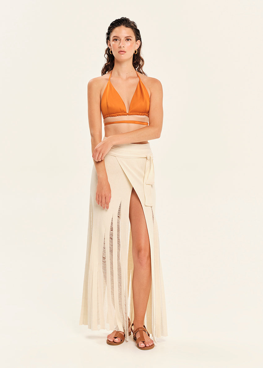 Woman wearing a mustard yellow bikini top and white skirt on a beige background
