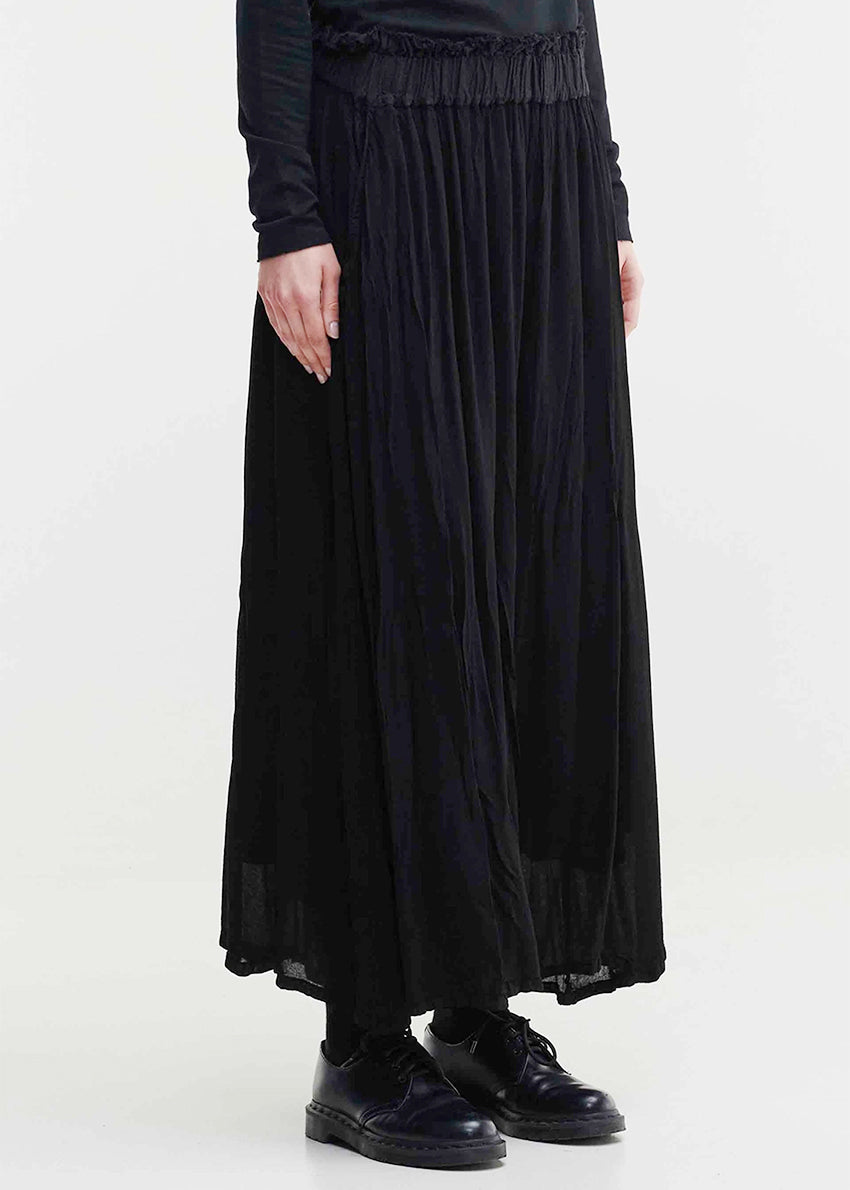 A person standing wearing Tanto Blues Pants, which are black color, with an elastic waist and side pockets. They are also wearing black shoes and a black top.