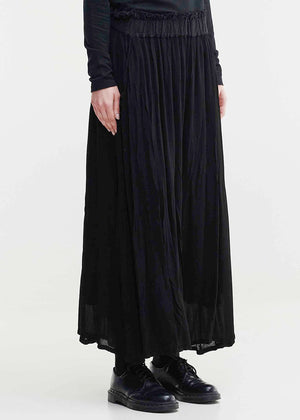 A person standing wearing Tanto Blues Pants, which are black color, with an elastic waist and side pockets. They are also wearing black shoes and a black top.