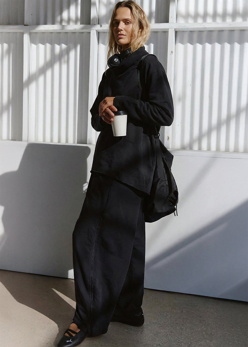 Person wearing a black outfit holding a white cup against a white wall with shadows.